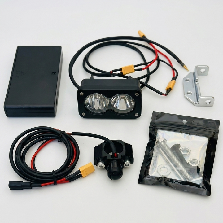 A dual LED off-road light kit with a black rectangular housing, two clear lenses, attached black wiring with yellow connectors, a black battery or power box, mounting hardware, and a bag of screws and washers, all arranged on a white background.
